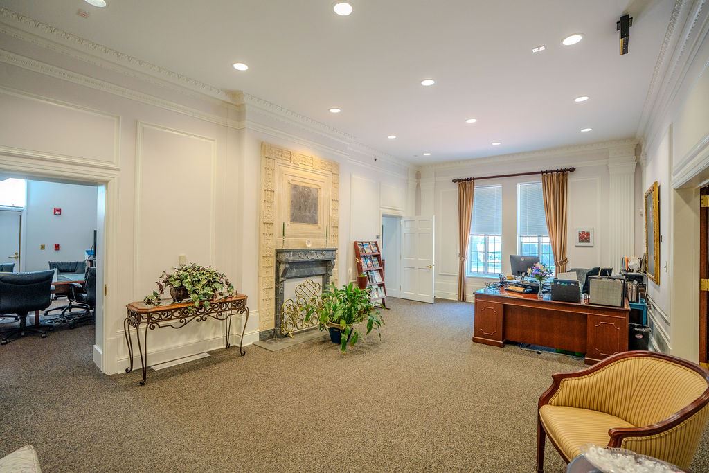 Large room with fireplace and mantel, desk, large windows, plants, and a couch.
